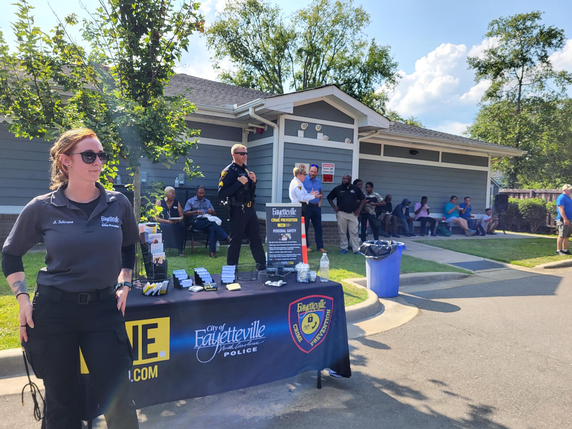 Outside a Crime Prevention Specialist standing at table. Officer standing near table and community members sitting near by.